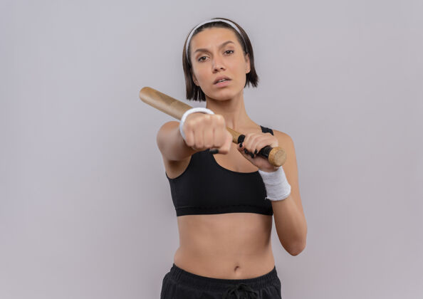 健身身穿运动服的年轻健身女士手持棒球棒 严肃的脸上露出拳头站在白色的墙上蝙蝠年轻女人