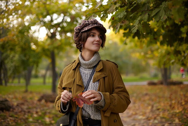 帽子时髦的棕色头发的年轻女士 bob发型 穿着时髦的保暖衣服 在温暖的秋日漫步在黄色的树上 手举树叶 面带微笑衣服女人鲍勃