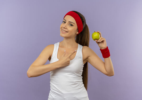 运动身穿运动服的年轻健身女士 头箍上拿着绿色的苹果 手指指着苹果 微笑着自信地站在灰色的墙上健康手指公民