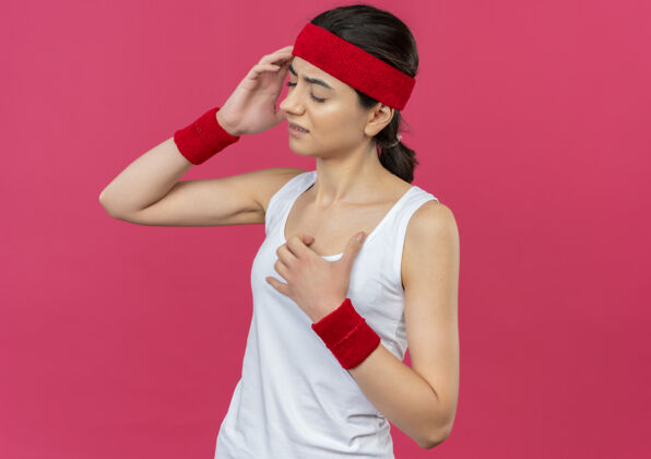 不适穿着运动服的年轻健身女 头戴头巾 看起来不舒服 站在粉红色的墙上摸着头痛的头市民头带运动