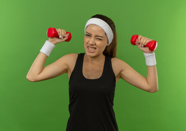 抱着身穿运动服 头箍 举着两个哑铃 表情烦躁地站在绿色墙壁上锻炼身体的年轻女子头带健身姿势