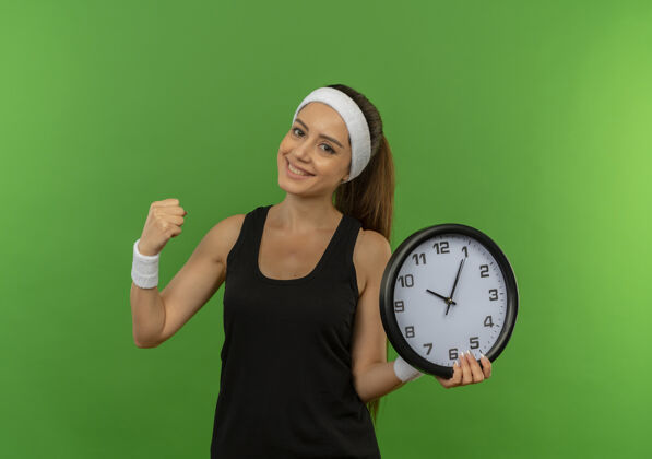 运动员身穿运动服的年轻健身女士 头戴花环 手持挂钟 握紧拳头 站在绿色的墙上 脸上洋溢着快乐和积极的微笑拳头运动年轻