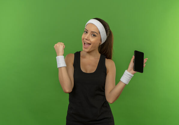人身穿运动服 头戴智能手机 握紧拳头的年轻健身女士站在绿色的墙壁上欢快地微笑着运动运动员女