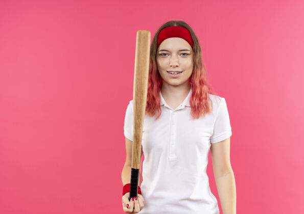 姿势戴着头巾的年轻运动型女子拿着球棒去打棒球 站在粉红色的墙上 看上去自信地微笑着人站着运动