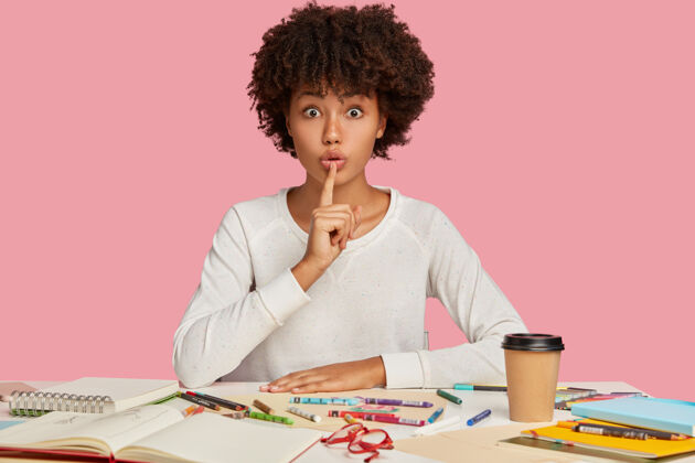 公共惊讶的深色皮肤的女性插画水平镜头保持食指在嘴上 演示沉默的手势 坐在桌面上用记事本 彩色铅笔 隔离在粉红色的墙上非洲女学生工人