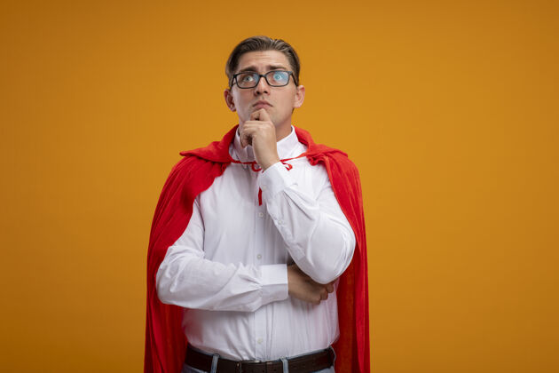 商人超级英雄商人穿着红色斗篷 戴着眼镜 站在橙色的背景下困惑地看着一边拼图超级英雄眼镜