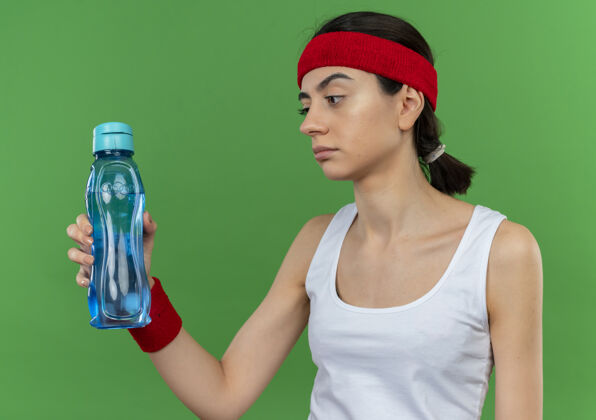 人身穿运动服的年轻健身女士戴着头巾 手里拿着一瓶水 站在绿色的墙上 带着怀疑的表情看着它 很不高兴健身不高兴站着
