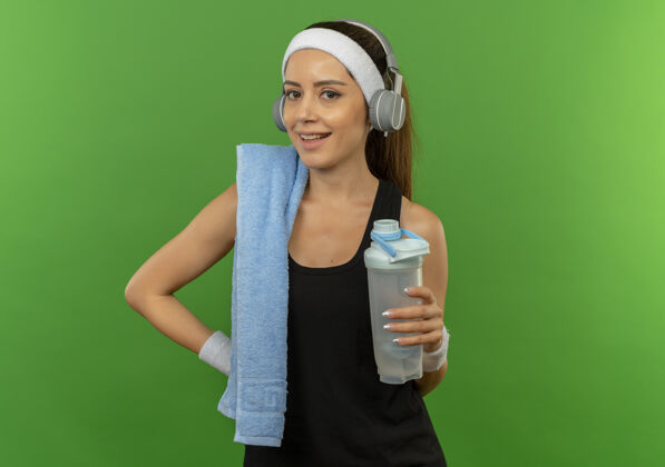 健康一位身穿运动服 头上戴着头巾 肩上扛着一瓶水的年轻健身女士站在绿色的墙上 脸上洋溢着欢快的笑容毛巾微笑人