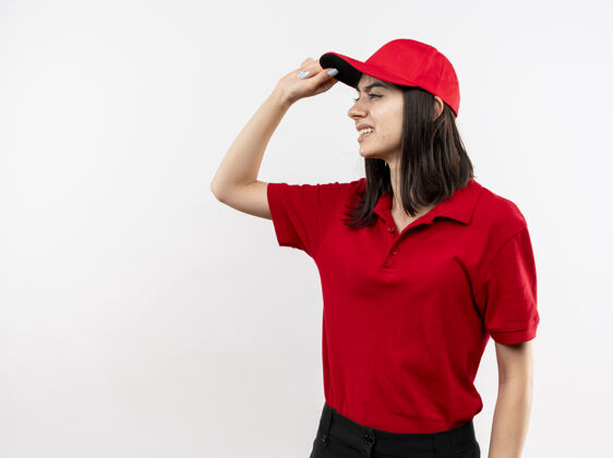 送货穿着红色制服 戴着帽子的年轻送货员一边看一边不高兴地碰了碰站在白色背景上的帽子站着年轻帽子