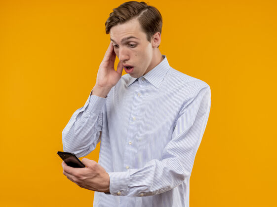 衬衫一个穿着白衬衫的年轻人拿着智能手机 站在橙色的背景下 看着它困惑和担心站着担心移动