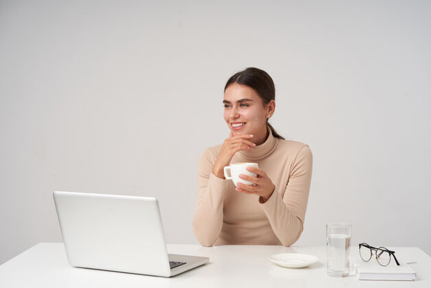 正式室内照片：迷人的年轻黑发女性 一边梦幻般地看着一边 面带微笑 一边喝着咖啡 一边用现代化的笔记本电脑在办公室工作化妆品头发眼镜
