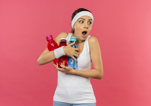 抱着穿着运动服的年轻健身女 头上戴着头巾 手里拿着几瓶水 站在粉红色的墙上惊讶地看着一边年轻人瓶子惊喜