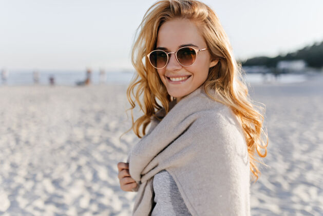 海景身着米色套装 戴着太阳镜的卷发年轻女子在海滩上微笑的快照水欢乐周末
