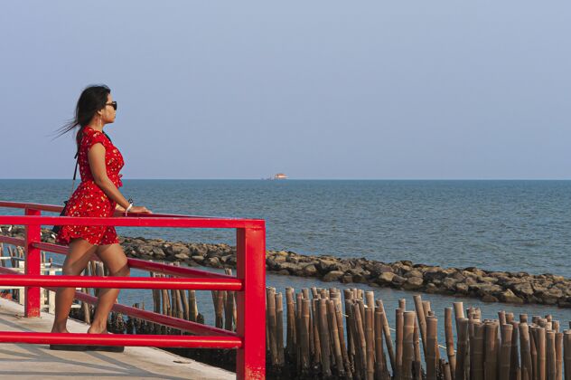阳光一个戴着太阳镜 穿着红色衣服的女人站在泰国的红色木板桥上模特晒黑海洋