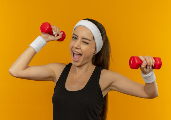 年轻身着运动服的年轻健身女士站在橘色的墙上 用两个哑铃做着练习 笑得很开心欢呼人运动