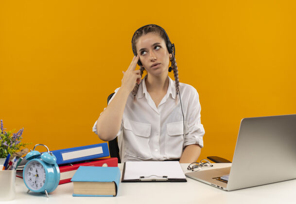 拼图一个留着辫子 穿着白衬衫 戴着耳机 拿着麦克风的年轻女学生一边看一边困惑 疲惫 无聊地坐在桌子旁 手里拿着笔记本电脑文件夹 剪贴板和橘色墙上的书姿势笔记本电脑坐着