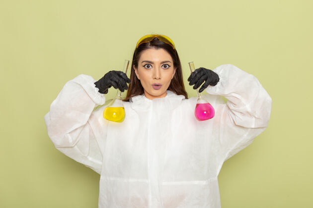 套头衫前视图穿着特殊防护服的女化学家拿着浅绿色表面上有溶液的烧瓶实验室化学家女