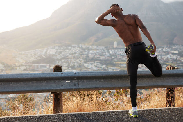 运动深色皮肤的男性站在后面 做伸展运动 抬腿 欣赏美丽的山景 有锻炼训练 穿运动鞋 站在沥青上健康和健身新鲜肌肉民族