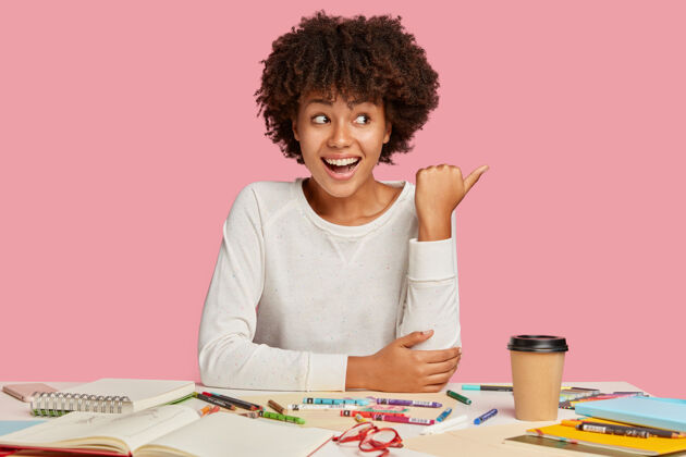 工作区快乐的黑人创意女士有积极的表情 用拇指指向一边 展示广告的自由空间 用螺旋笔记本和蜡笔在工作场所摆姿势 隔离在粉红色的墙上办公桌工作蜡笔