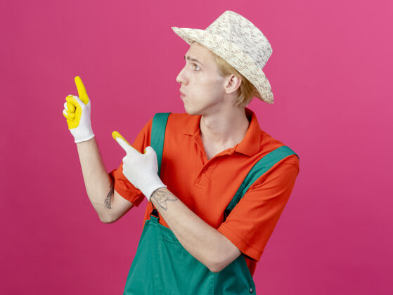 花园年轻的园丁穿着连体衣戴着帽子戴着橡胶手套用食指指着年轻人连身衣男人