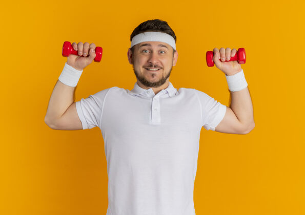 自信身穿白衬衫 头箍 举着哑铃的年轻健身男子站在橙色的墙上自信地微笑着运动男性站立
