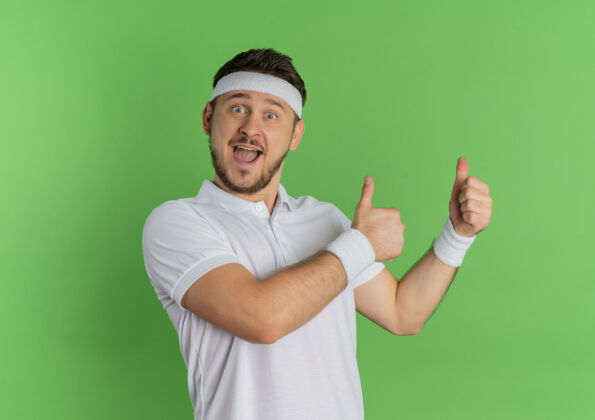 运动员身穿白衬衫 头箍的年轻健身男子站在绿色的墙上 面带微笑 高高兴兴地竖起大拇指衬衫表演运动装