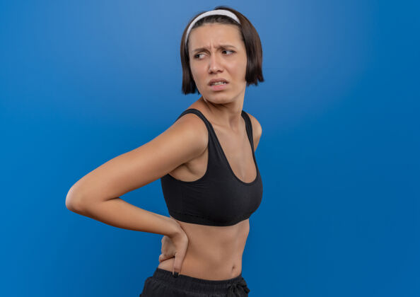 焦虑穿着运动服的年轻健身女士站在蓝色的墙壁上 摸着她的背 困惑地看着一边 非常焦虑人姿势困惑