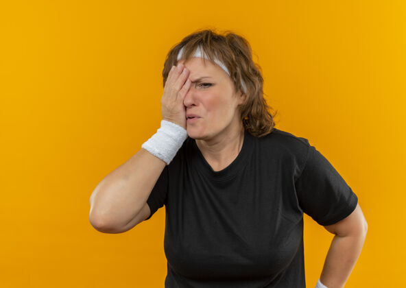 运动装中年运动女性 身穿黑色t恤 戴着头带 看起来很累 一只眼睛被遮住 胳膊站在橙色的墙上女人老年姿势