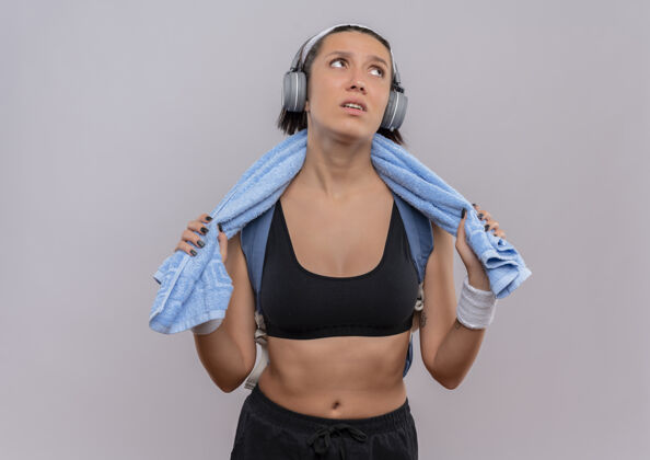 毛巾年轻的健身女士头戴耳机 脖子上套着毛巾 站在白色的墙边 疲惫而无聊地看着一边脖子无聊旁边