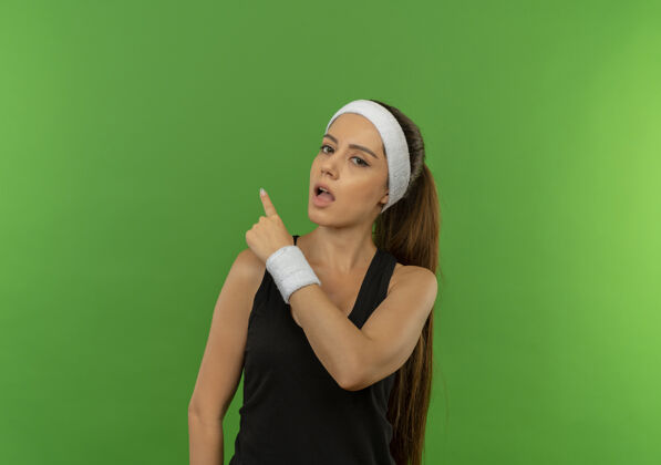 壁板戴着头巾的年轻健身女士困惑地用手指着站在绿墙上的一边看运动人