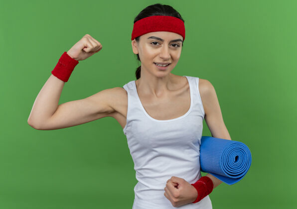 女人身穿运动服的年轻健身女士 头戴瑜伽垫 自信地举起拳头 站在绿色墙壁上的赢家理念健身姿势运动装