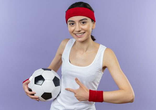 微笑身穿运动服的年轻健身女 头戴足球带 食指指着足球 微笑着站在紫色的墙上指着女人手指