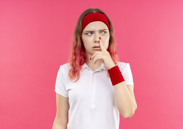 运动戴着头巾的年轻运动女郎站在粉红色的墙上 一边看一边抚摸着鼻子 脸上带着沉思的表情女人市动员
