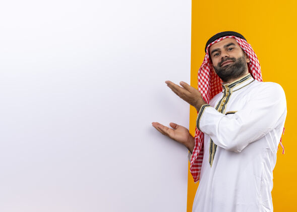 阿拉伯语身着传统服装的阿拉伯商人手举空白广告牌站在橙色的墙上介绍站传统
