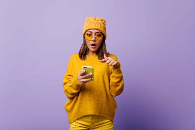 服装穿着黄色裤子和大号毛衣的女士惊呆了 她的智能手机里有一封奇怪的电子邮件紫色墙壁上的情感模特的明亮肖像现代模特橙色