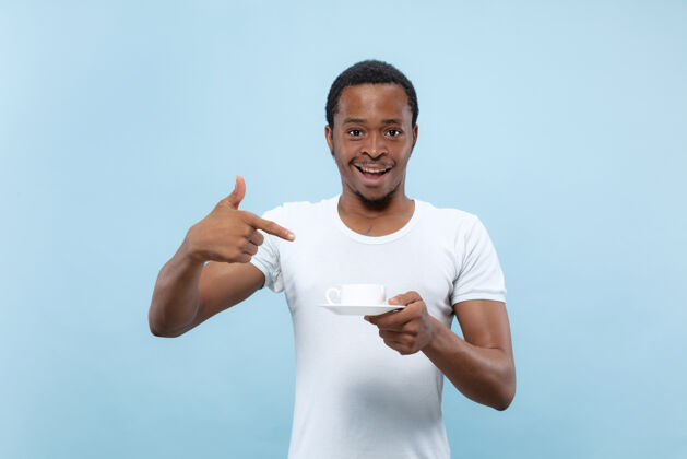 人半身特写 蓝色背景 身穿白衬衫的非洲裔美国年轻男子人类情感 面部表情 广告 销售 概念享受 喝咖啡 微笑指指点点就业胡须事业