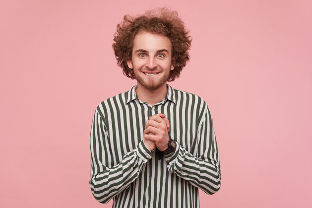 年轻兴奋的年轻绿眼睛胡须红发男子穿着休闲服折叠举起的手和咬内衣 而站在粉红色的墙上条纹衬衫折叠发型漂亮