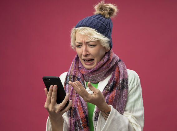 感觉心烦意乱的生病的不健康的女人 短头发 戴着暖和的围巾和帽子 感觉不舒服 压力和沮丧看着她站在粉色背景下的智能手机屏幕生病手机头发