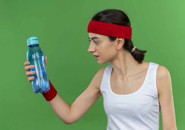 人穿着运动服的年轻健身女 戴着头巾 手里拿着一瓶水 站在绿色的墙上困惑地看着它头带运动装抱着