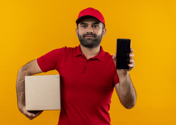 看着留着胡须的送货员穿着红色制服 戴着帽子 拿着一个盒子包 站在橙色的墙上 智能手机看起来很自信帽子制服包装