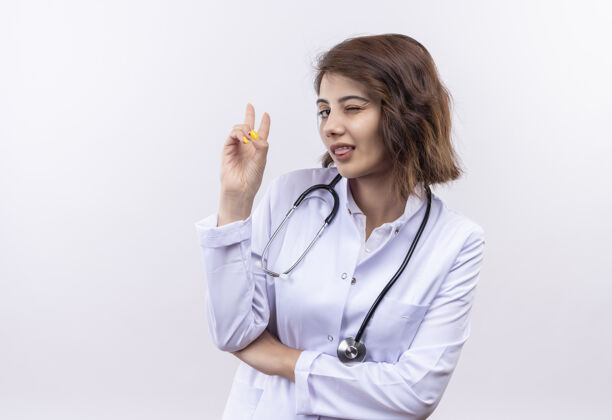 女人身穿白大褂的年轻女医生用听诊器眨眼 显示胜利的标志 伸出舌头站在白墙上眨眼外套胜利