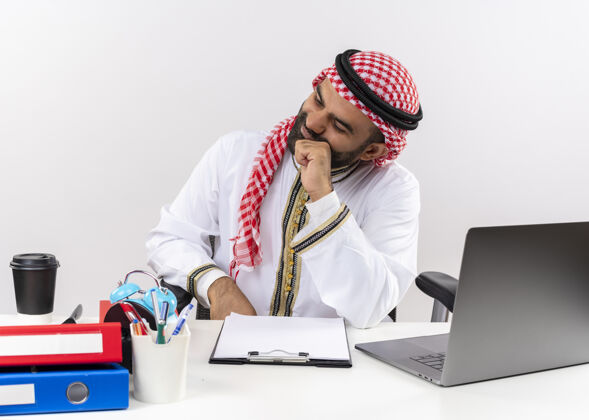 笔记本电脑身着传统服装的阿拉伯商人坐在桌旁 拿着笔记本电脑 神情沉思 困惑地在办公室工作沉思拼图电脑