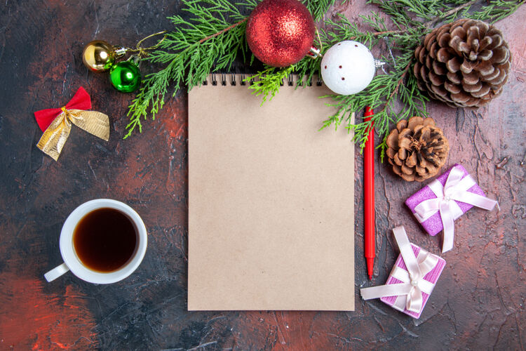 顶部顶视图红色钢笔笔记本松树树枝圣诞树球玩具和礼物一杯茶在暗红色的表面背景笔记本茶