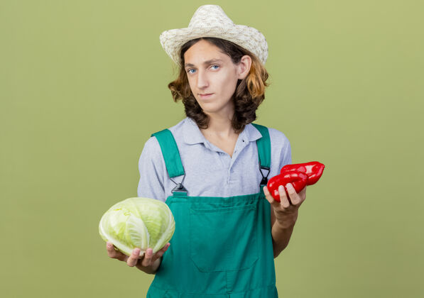 卷心菜年轻的园丁 穿着连体衣 戴着帽子 手里拿着卷心菜和红甜椒花园红色站着