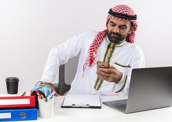 桌子身着传统服装的阿拉伯商人坐在明亮的办公桌旁 用智能手机发短信信息商人工作