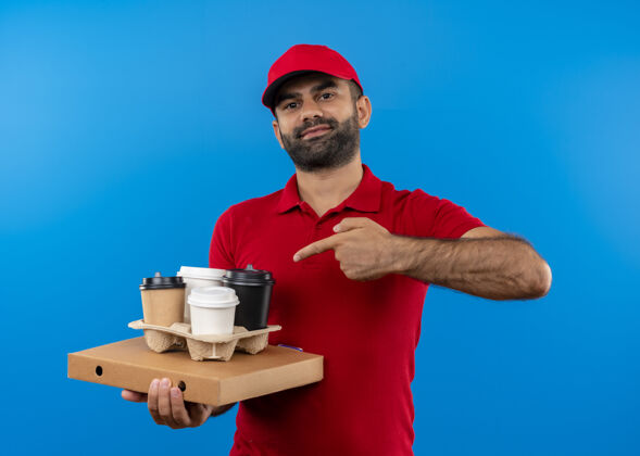 站着留着胡须的送货员身穿红色制服 戴着帽子 手里拿着披萨盒和咖啡杯 手指指着他们 站在蓝色的墙上自信地微笑着送货帽子咖啡