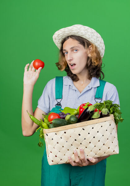 帽子年轻的园丁穿着连体衣戴着帽子拿着装满新鲜蔬菜的箱子年轻男人穿