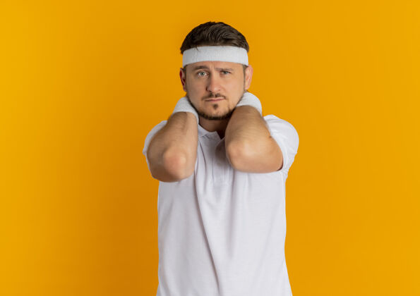 运动身穿白衬衫 头箍 脸上带着悲伤表情的年轻健身男子站在橙色的墙上望着前方健康运动小伙子