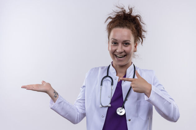 戴着年轻女医生身穿白色外套 手持听诊器 用食指指着站在白墙上的一侧站着手指胳膊