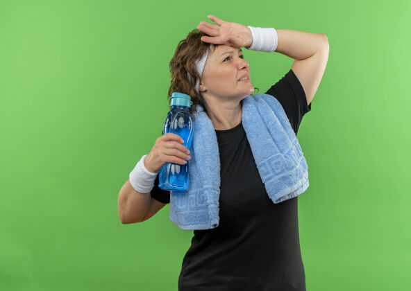 毛巾一位中年运动女性 身穿黑色t恤 脖子上戴着头带和毛巾 站在绿色的墙上 手里拿着一瓶水 看上去很疲惫女人头带抱着